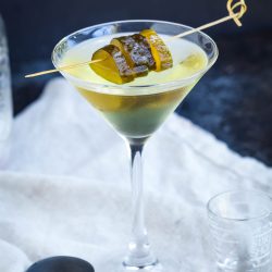 A martini glass filled with a clear yellow-green cocktail, garnished with pickle slices on a wooden skewer, sits on a white cloth with bar tools nearby.