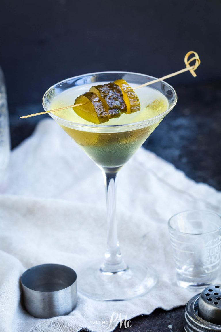 A martini glass filled with a clear yellow-green cocktail, garnished with pickle slices on a wooden skewer, sits on a white cloth with bar tools nearby.