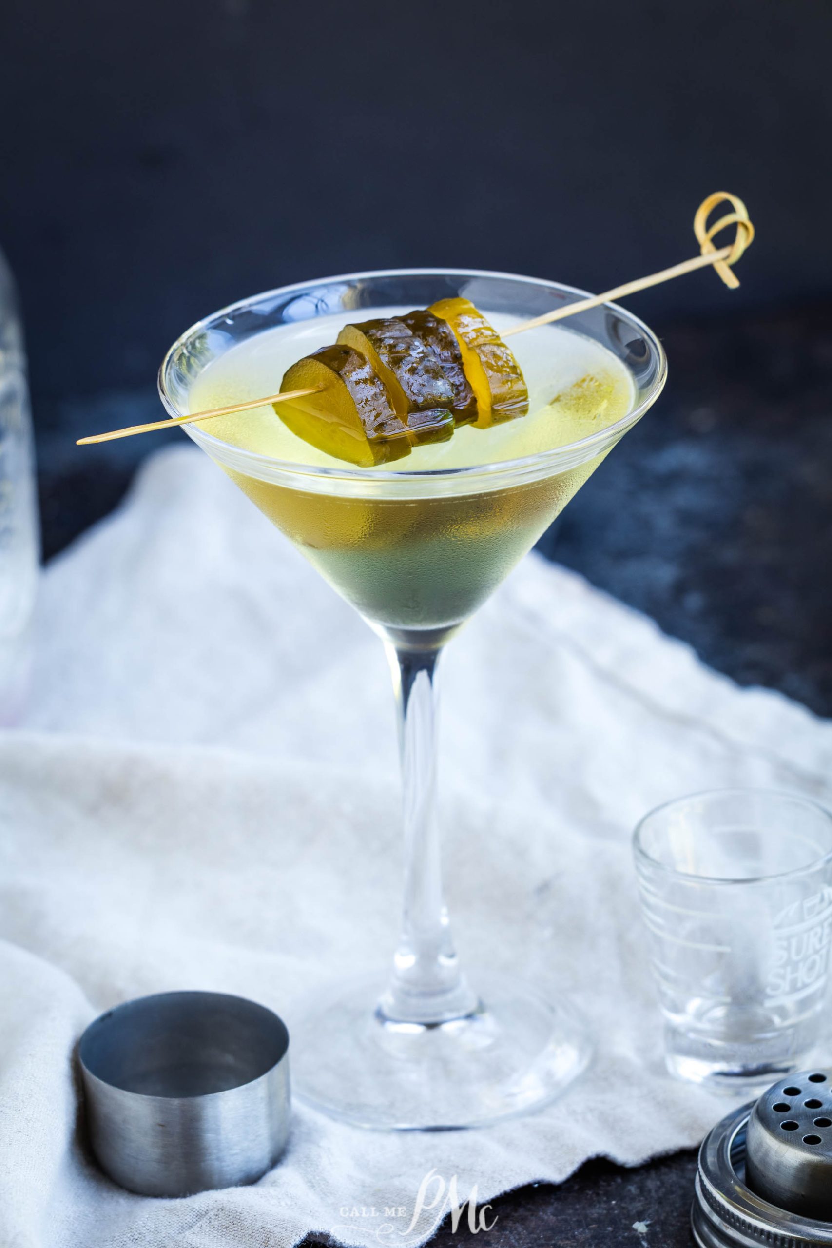A glass filled with a clear yellow-green cocktail, garnished with wooden skewer, sits on a white cloth with bar tools nearby.
