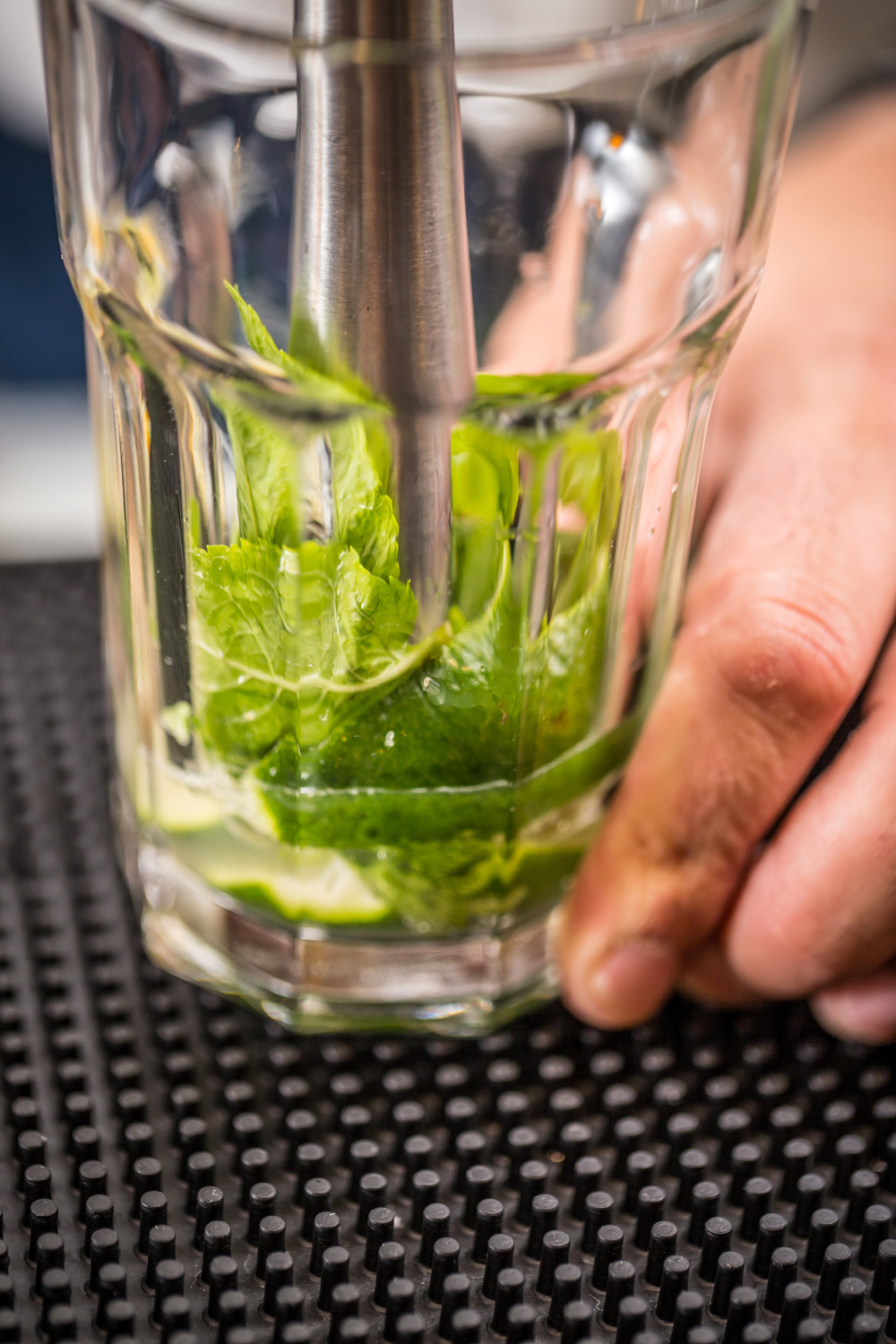 A close-up of a person muddling fresh mint leaves and lime in a glass with a metal muddler, preparing a cocktail on a black textured bar mat.