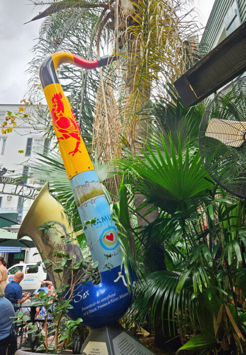 A large, colorful saxophone sculpture with various designs stands among lush green plants outdoors; people and buildings are visible in the background.