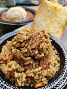 A close-up of a bowl filled with jambalaya, featuring rice, sausage, and vegetables, with a slice of garlic bread in the background. Another bowl with mashed potatoes and beans is visible behind.