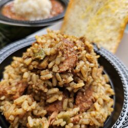 A close-up of a bowl filled with jambalaya, featuring rice, sausage, and vegetables, with a slice of garlic bread in the background. Another bowl with mashed potatoes and beans is visible behind.