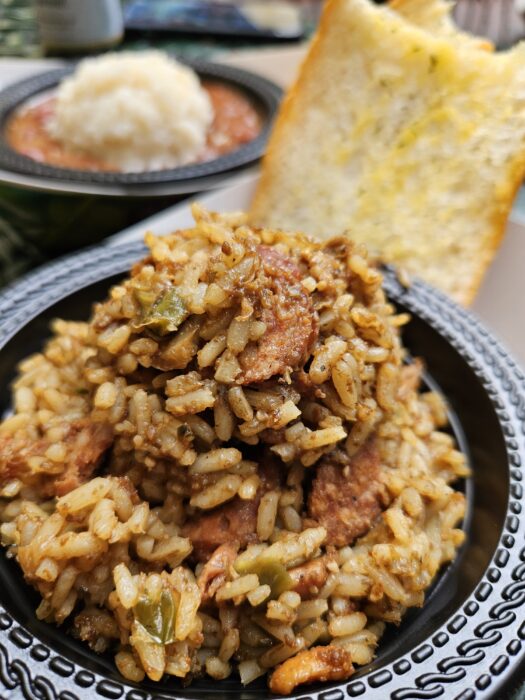 A close-up of a bowl filled with jambalaya, featuring rice, sausage, and vegetables, with a slice of garlic bread in the background. Another bowl with mashed potatoes and beans is visible behind.