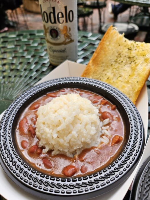 A bowl of red beans with a mound of white rice in the center, served with a slice of garlic bread on the side. A bottle of Modelo beer is in the background on a green, textured table.
