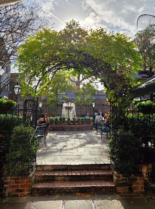 A garden arch covered in green leaves frames a courtyard with brick steps, outdoor tables, lush plants, and a central fountain. People sit around, enjoying the sunny, peaceful atmosphere.