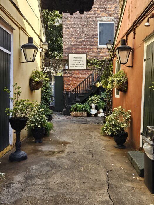 A narrow, sunlit alleyway with yellow and orange walls, black lanterns, potted plants, and a brick building at the end with a welcome sign and a staircase leading up to a door.
