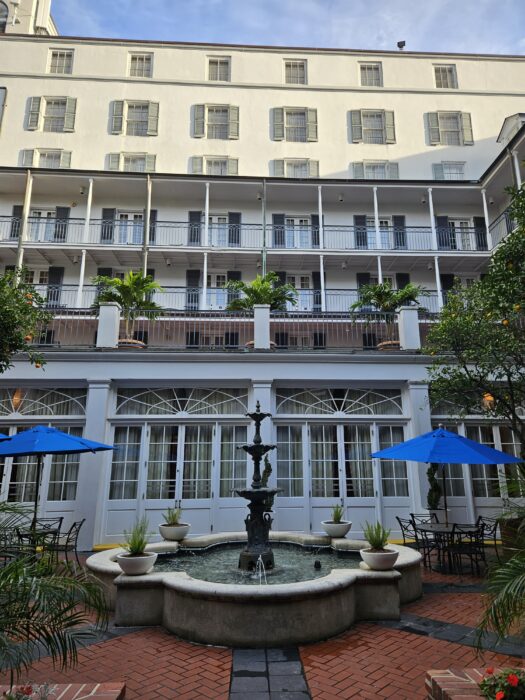 A courtyard with a central fountain, surrounded by potted plants, outdoor tables, and blue umbrellas. Behind it is a multi-story white building with balconies and shuttered windows.