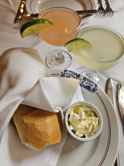 A white plate with a roll of bread wrapped in a napkin, a small dish of whipped butter, and two cocktails garnished with lime wedges on a white tablecloth. Silverware is visible in the background.