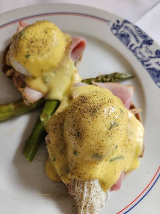 Two eggs Benedict on a white plate with blue trim, topped with creamy hollandaise sauce and black pepper, served on slices of ham with asparagus spears underneath.