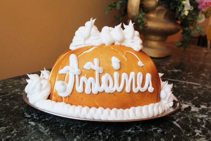 A dome-shaped cake with golden-brown crust, decorated with white frosting that spells “Antoine’s,” sits on a marble surface. Whipped cream details adorn the cake’s top and base. A decorative vase is blurred in the background.