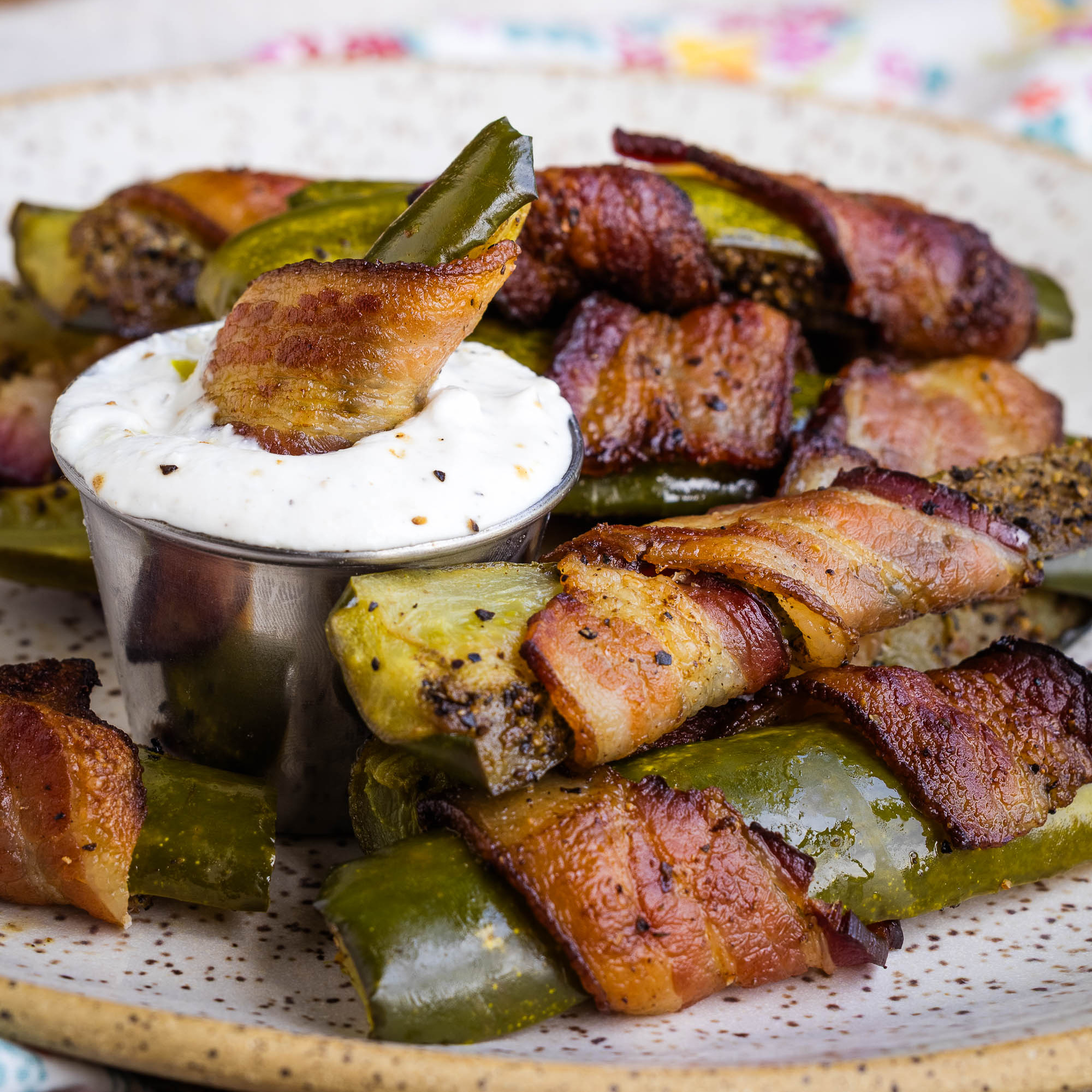 Bacon-wrapped jalapeño poppers served on a plate, with one popper dipped in a small metal cup of creamy white sauce. The poppers are roasted and visibly seasoned.