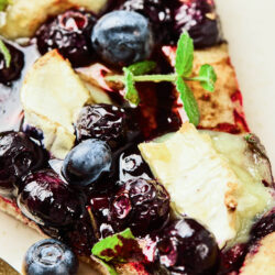 Close-up of a blueberry goat cheese flatbread slice topped with fresh blueberries, melted cheese, sprigs of mint, and a golden, crispy crust. A gold fork rests beside the flatbread on a light-colored plate.
