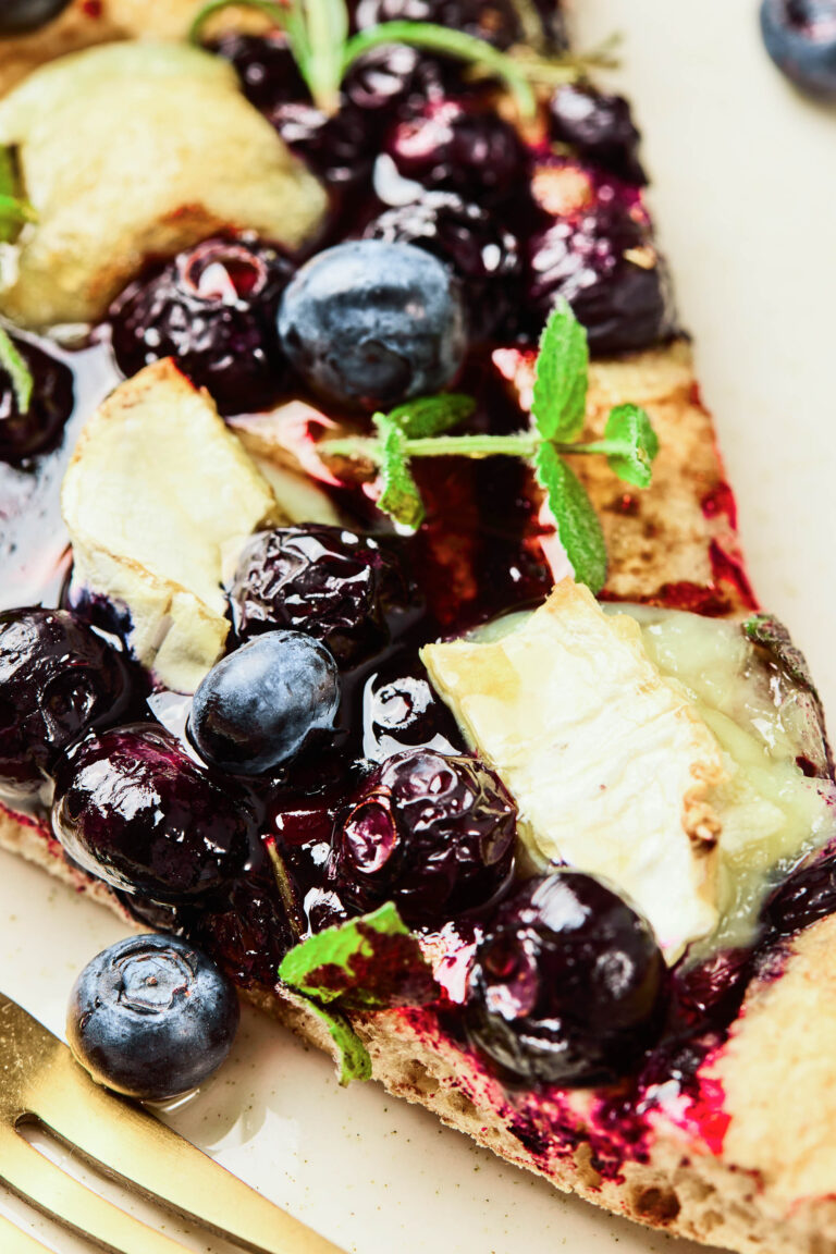 Close-up of a blueberry goat cheese flatbread slice topped with fresh blueberries, melted cheese, sprigs of mint, and a golden, crispy crust. A gold fork rests beside the flatbread on a light-colored plate.