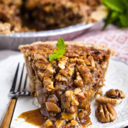 A slice of pecan pie on a white plate, with caramel filling oozing out. A fork, pecans, and a mint garnish are on the plate. The rest of the pie and a sprig of mint are in the background.