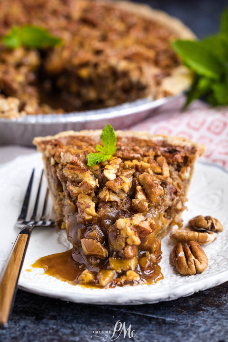 A slice of pecan pie on a white plate, with caramel filling oozing out. A fork, pecans, and a mint garnish are on the plate. The rest of the pie and a sprig of mint are in the background.