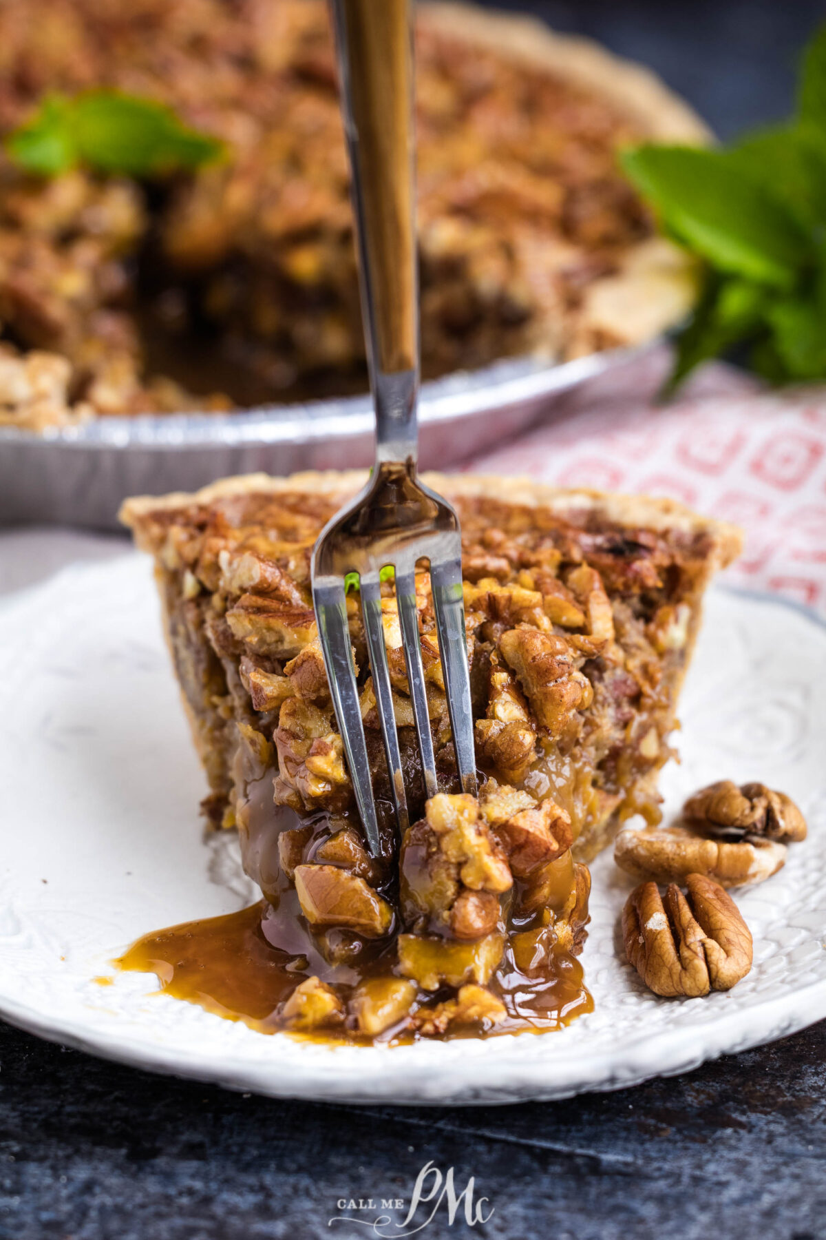 A slice of pecan pie on a white plate with a fork inserted, caramel filling oozing out. Whole pecans are next to the pie, and the rest of the pie is visible in the background.