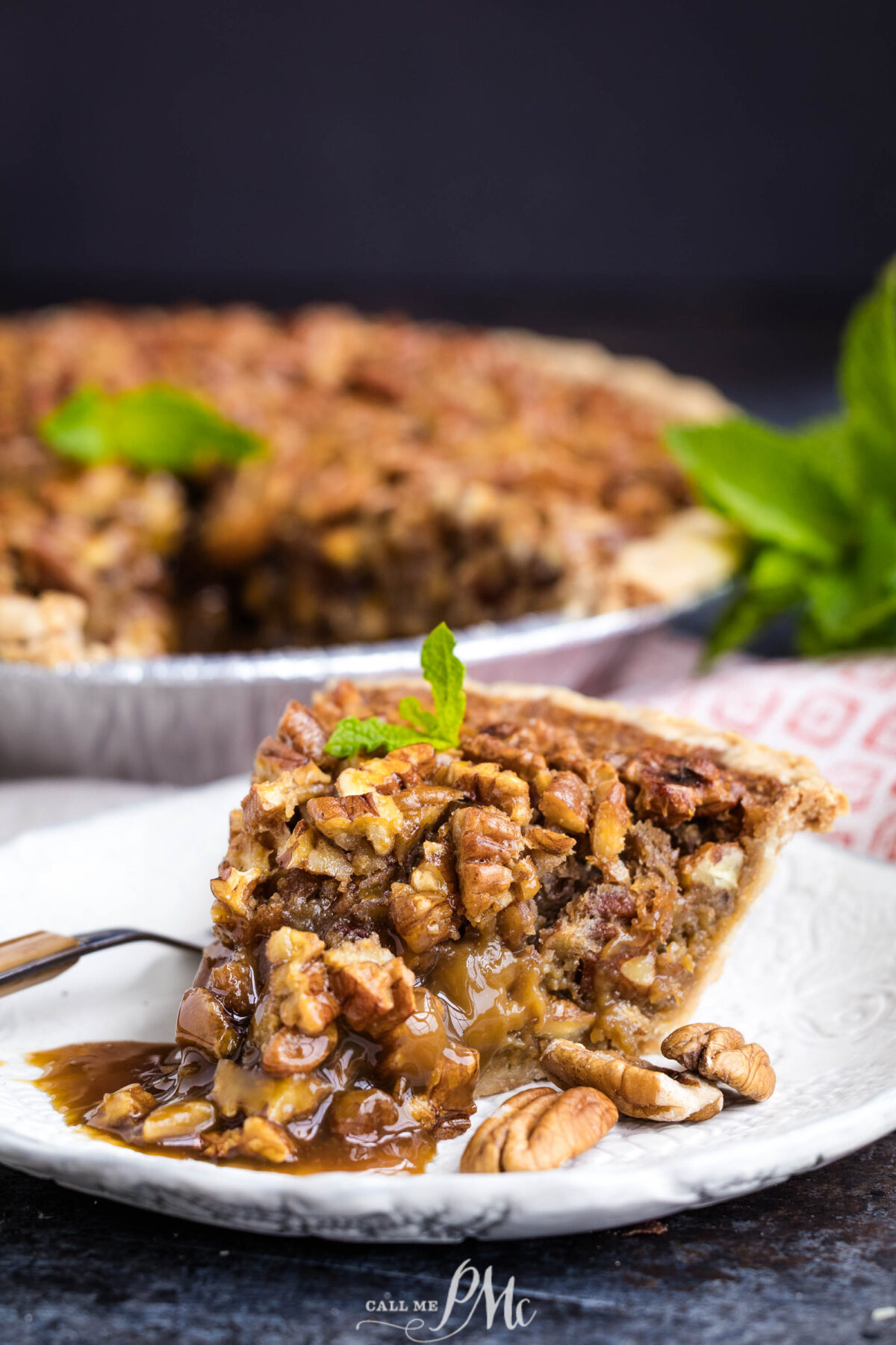 A close-up of a slice of pecan pie on a white plate, garnished with a mint sprig. The filling is gooey and rich, with whole pecans visible. The whole pie is in the background, slightly out of focus.