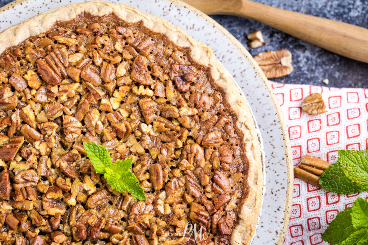 A close-up of a Southern bourbon pecan pie with garnished with fresh mint leaves, placed on a speckled plate. A red-and-white towel, pecans, and a wooden spoon are nearby on a textured surface.