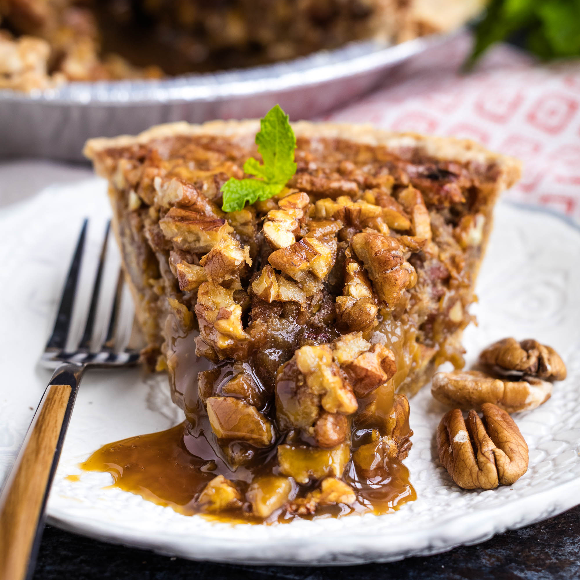 A slice of pecan pie with a gooey caramel filling sits on a white plate, garnished with a sprig of mint, alongside a fork and a few pecans.