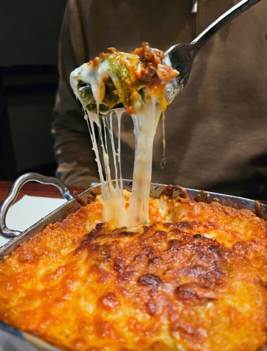 A fork lifts a cheesy, gooey portion of baked lasagna from a pan, with melted cheese stretching from the dish. A person in a brown top is partially visible in the background.