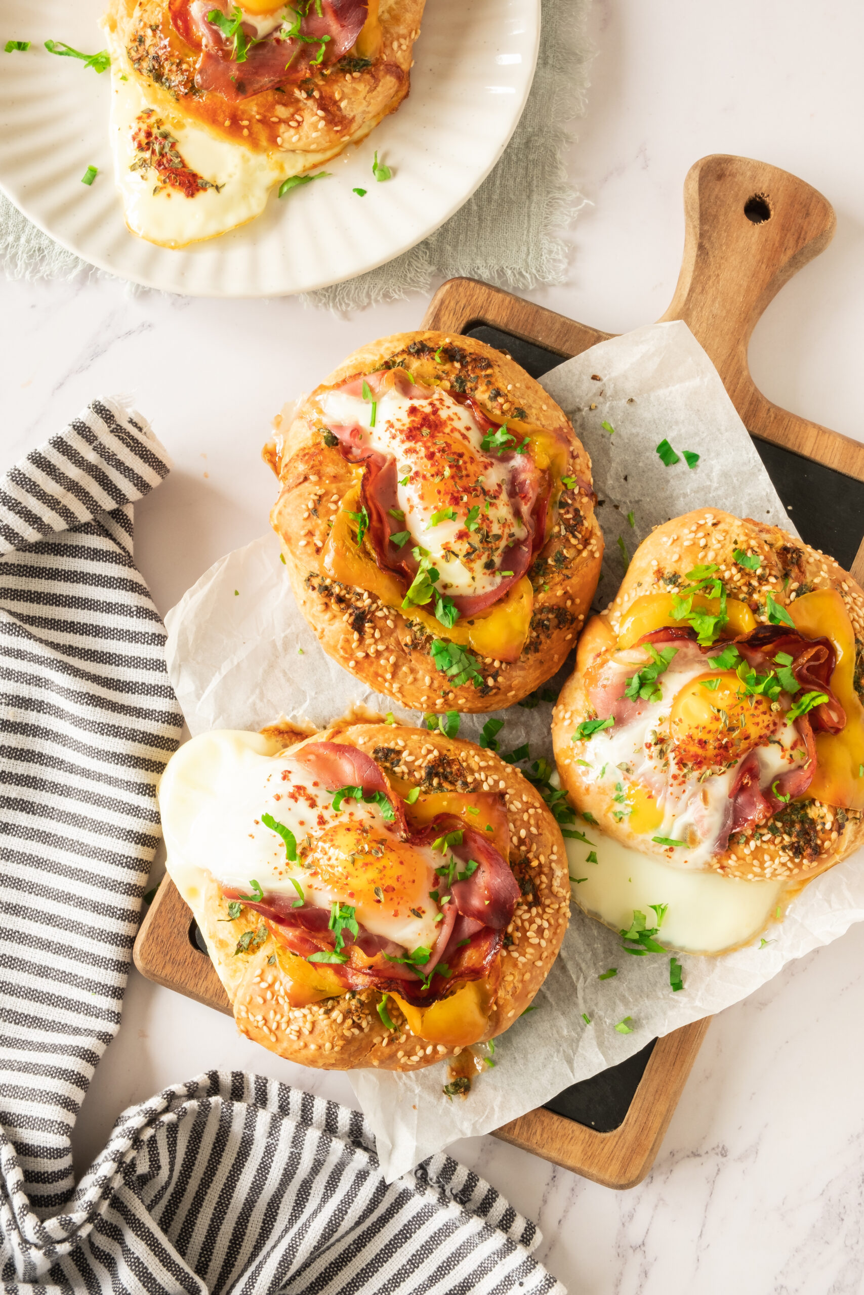 Three open-faced sandwiches, reminiscent of a bagel with egg prosciutto and garlic butter, feature melted cheese, ham, and herbs on a wooden board lined with parchment. One more sandwich rests on a plate by a striped napkin atop white marble.