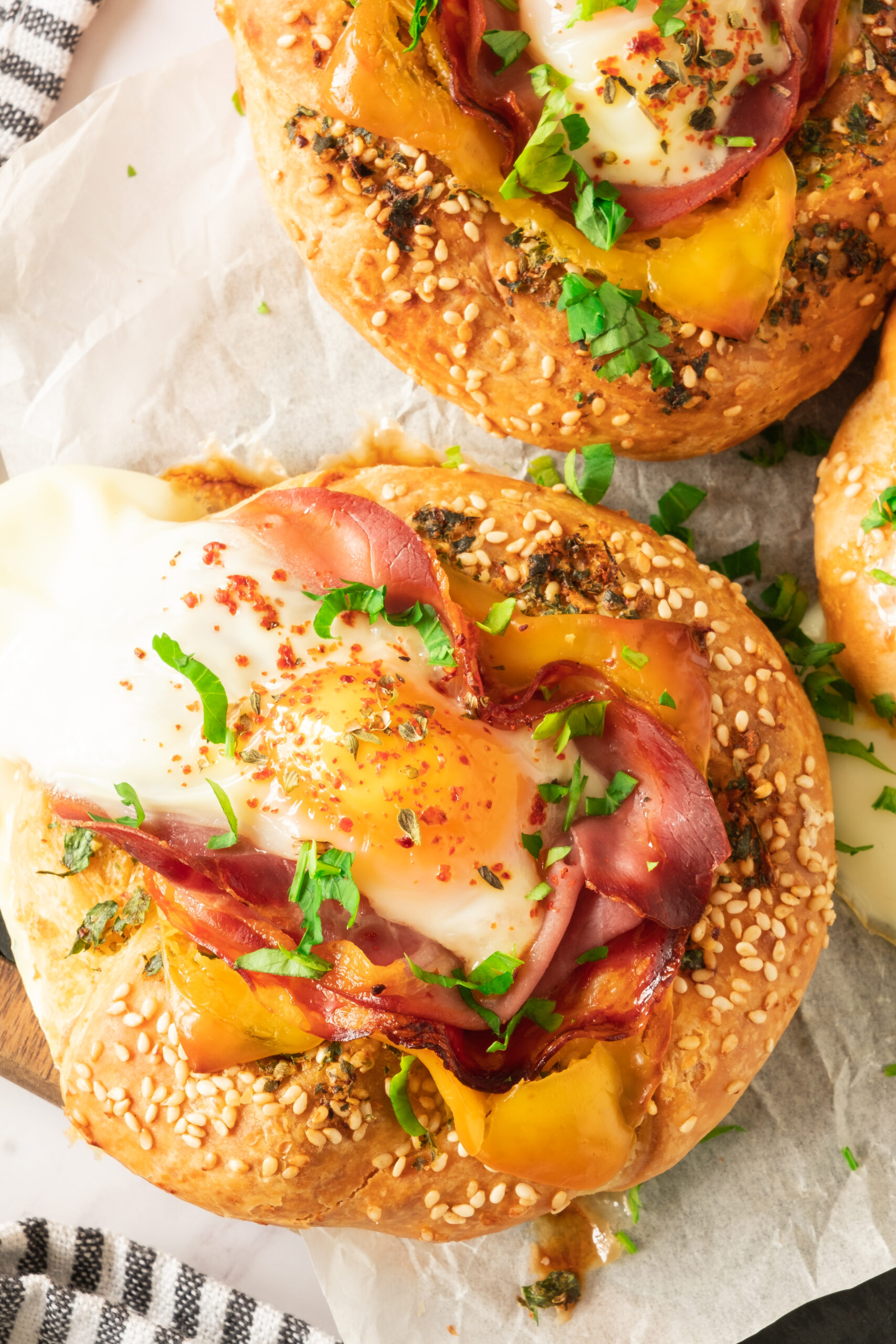 Breakfast sandwich topped with melted cheese and garnished with chopped herbs and chili flakes, placed on parchment paper.