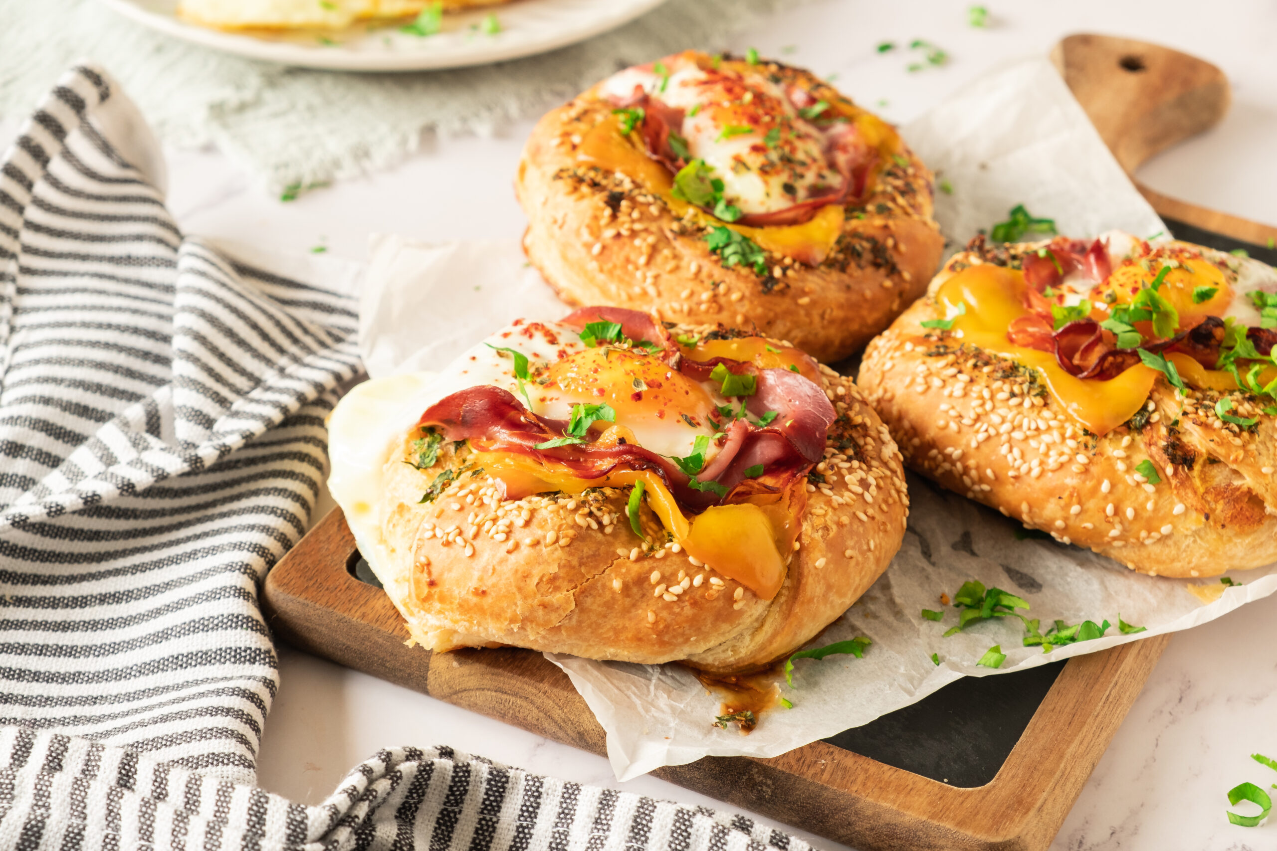 Three sesame seed bread rolls, reminiscent of a bagel with egg prosciutto and garlic butter, are topped with eggs, ham, bell peppers, and herbs on parchment paper. Fresh parsley is sprinkled on top, with a striped cloth lying nearby.