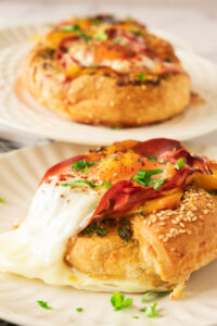 Two breakfast pastries, inspired by a Bagel with Egg Prosciutto and Garlic Butter, are topped with melted cheese, baked egg, prosciutto, herbs, and sesame seeds—served on white plates, one in focus and another blurred in the background.