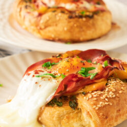Two breakfast pastries, inspired by a Bagel with Egg Prosciutto and Garlic Butter, are topped with melted cheese, baked egg, prosciutto, herbs, and sesame seeds—served on white plates, one in focus and another blurred in the background.