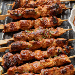 Firecracker Chicken Skewers with a golden-brown, slightly charred surface are arranged on a grill pan with lemon wedges on the side. A small dish of sauce is partially visible.