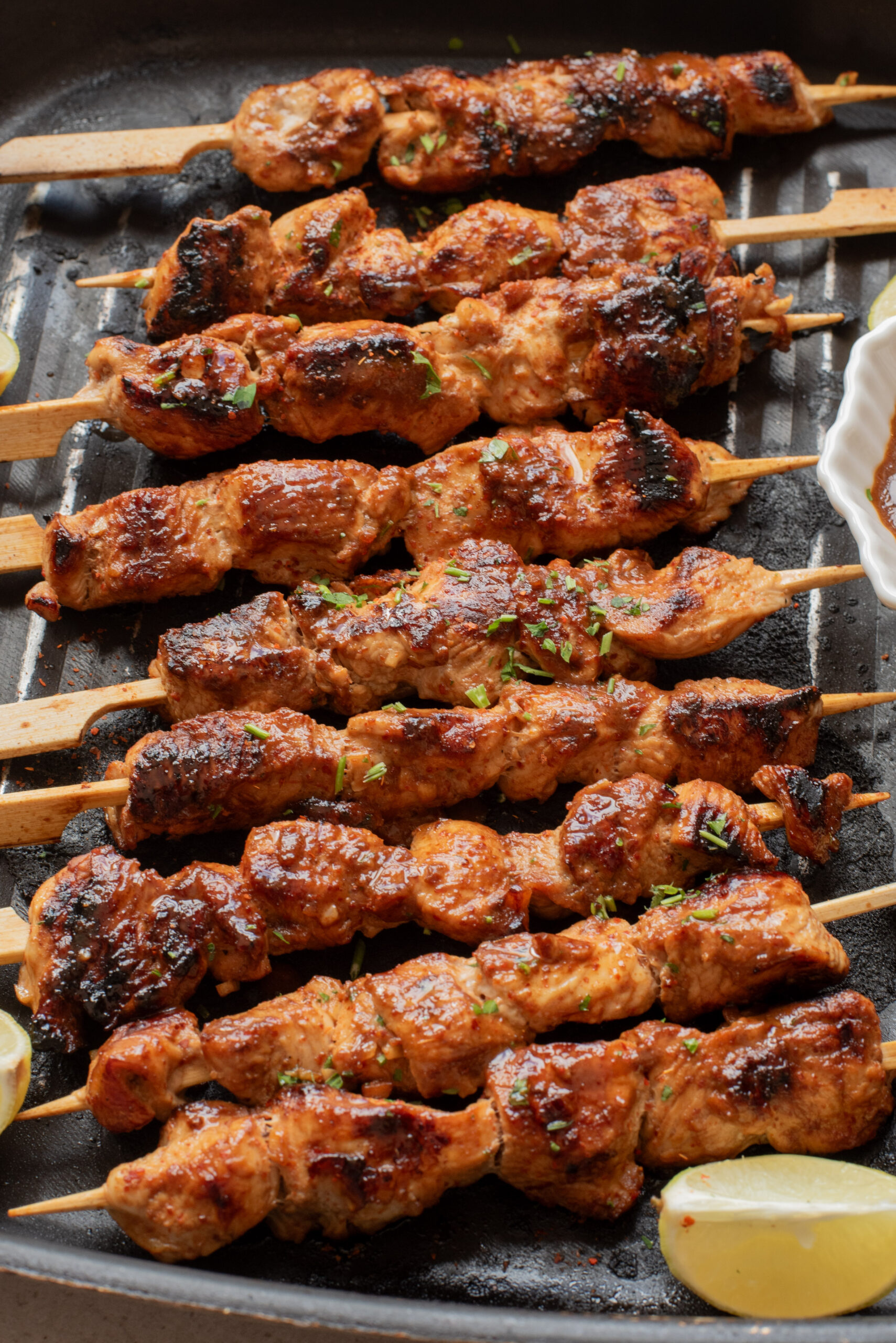 Kabobs with a golden-brown, slightly charred surface are arranged on a grill pan with lemon wedges on the side. A small dish of sauce is partially visible.