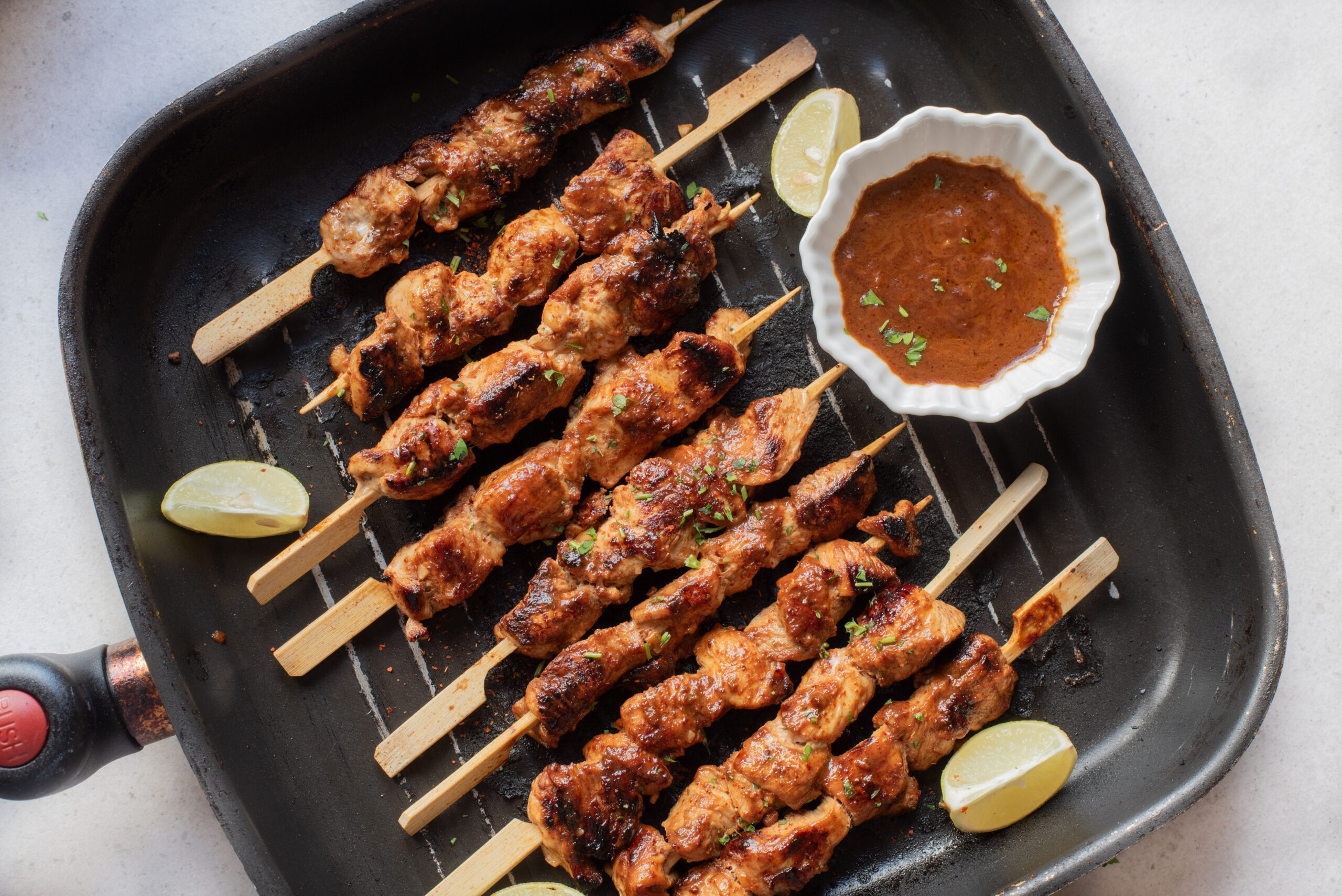 Firecracker Chicken Skewers are arranged in a grill pan with three lime wedges and a small bowl of brown dipping sauce, garnished with chopped herbs.