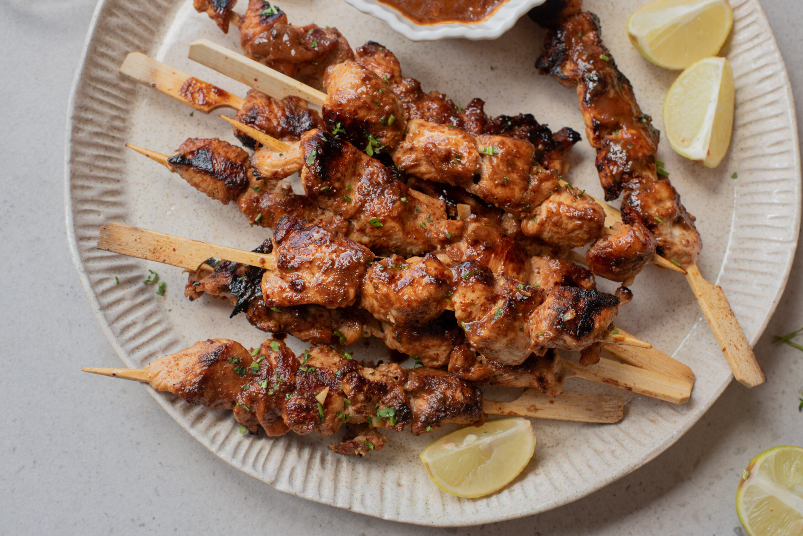 Firecracker Chicken Skewers served on a plate with fresh lemon wedges and a small bowl of dipping sauce nearby. The chicken is slightly charred, garnished with herbs for a bold, flavorful bite.