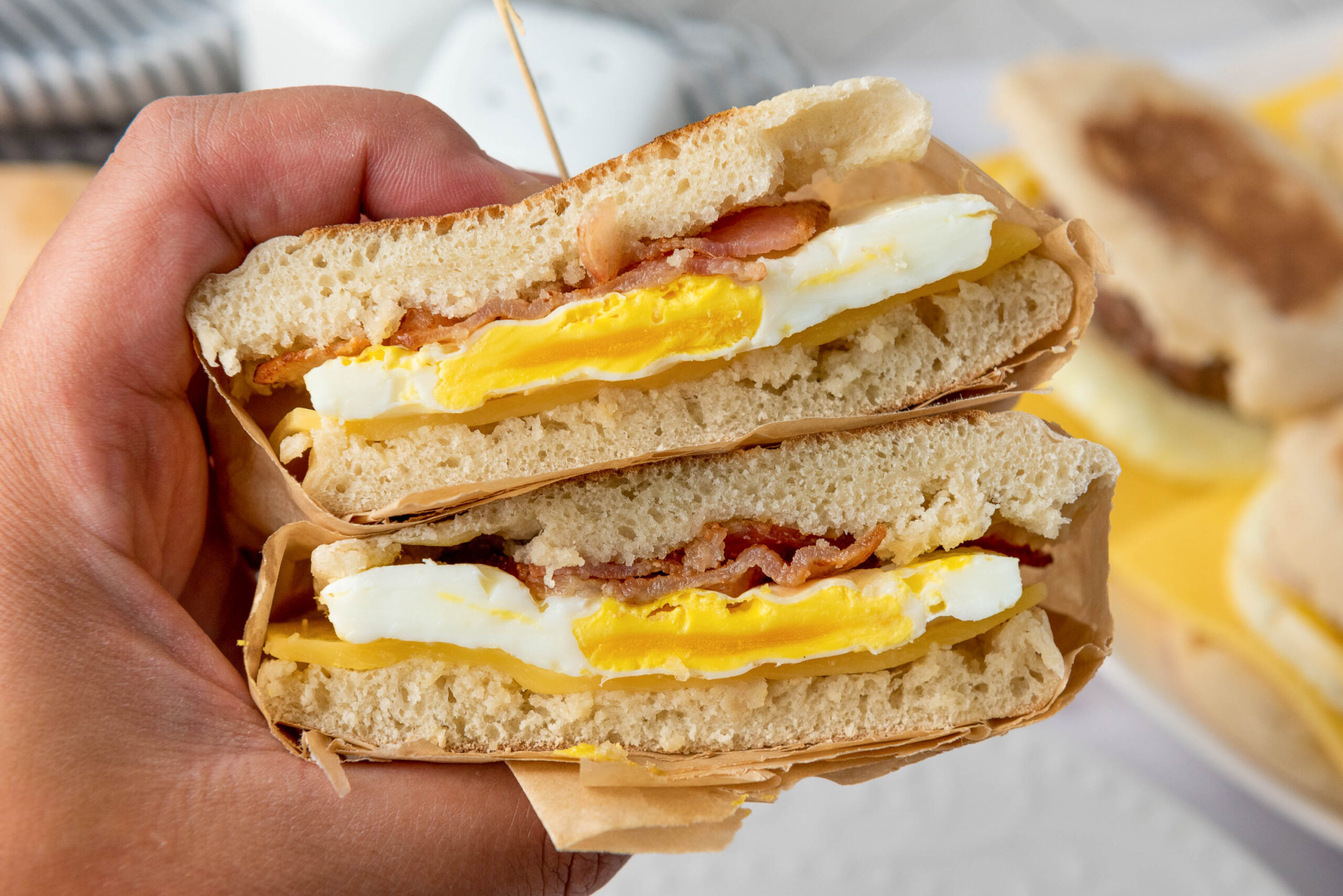 A hand holds a breakfast sandwich cut in half, showing layers of egg, bacon, and cheese between halves of an English muffin—perfect for easy freezer breakfast sandwiches. Another sandwich is blurred in the background.