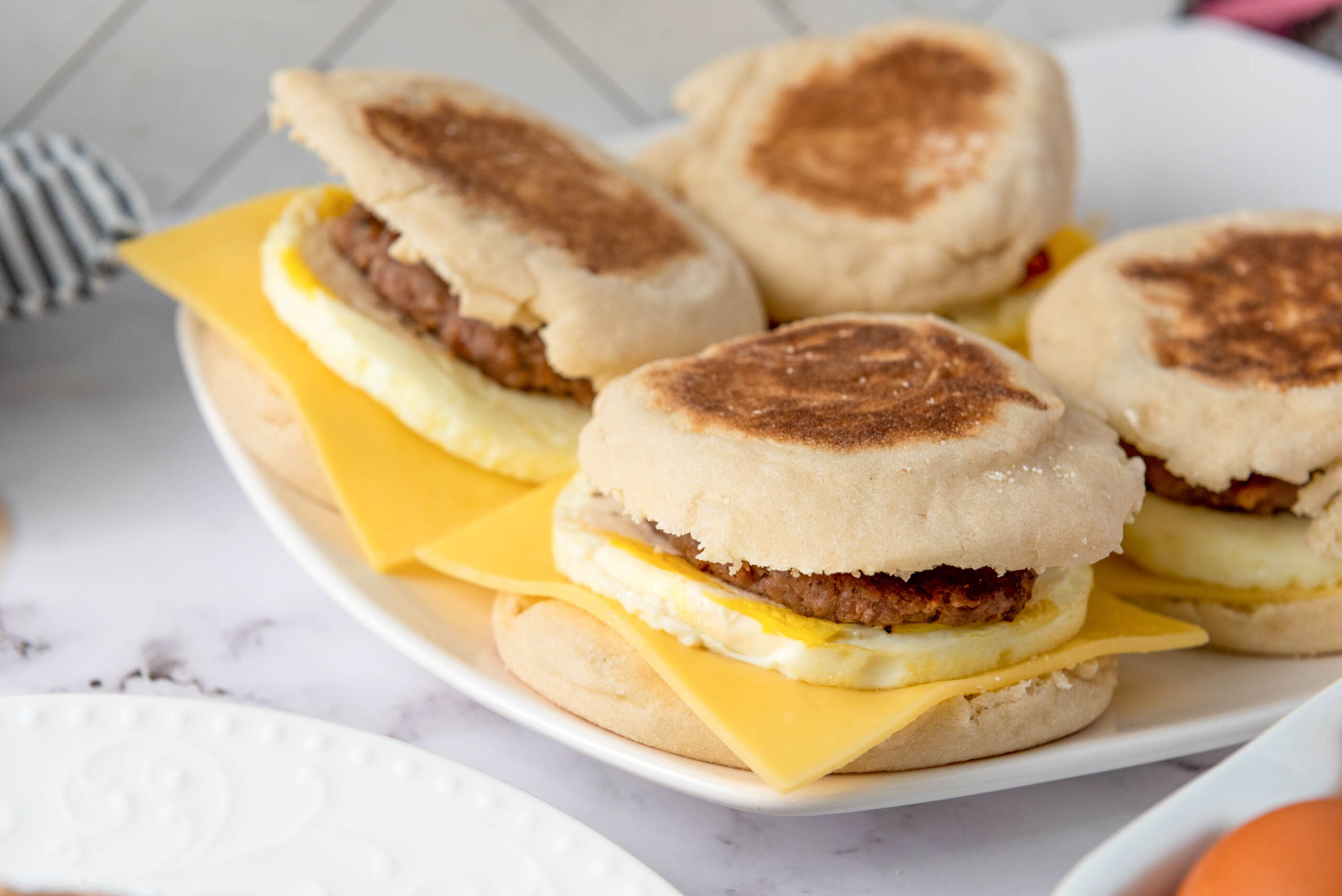 A plate of freezer breakfast sandwiches, each made with an English muffin, a slice of cheddar cheese, a cooked egg, and a sausage patty. The sandwiches are arranged closely together on a white plate.