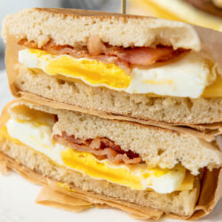 A close-up of a breakfast sandwich cut in half, showing layers of scrambled eggs, crispy bacon, and bread—perfect inspiration for homemade freezer breakfast sandwiches—served on parchment paper on a white plate.