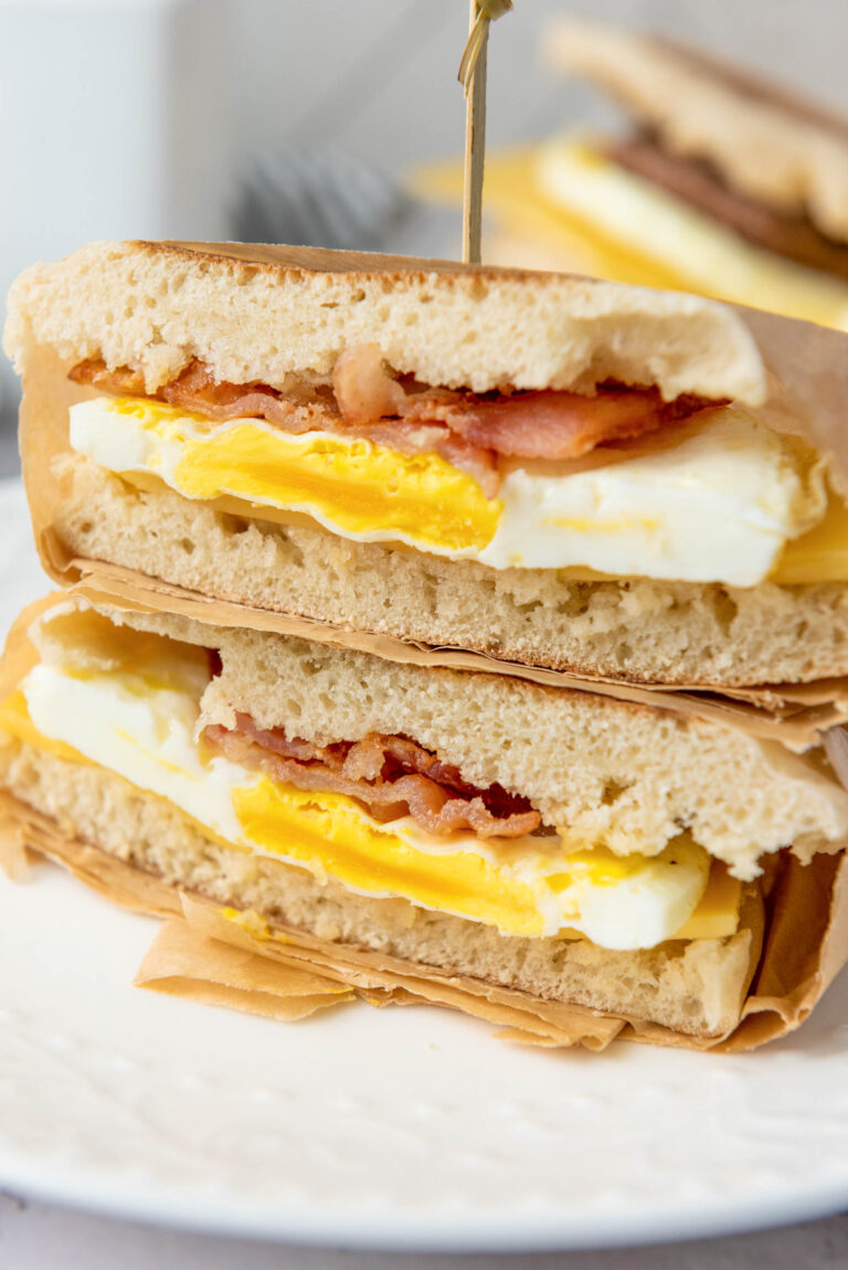 A close-up of a breakfast sandwich cut in half, showing layers of scrambled eggs, crispy bacon, and bread—perfect inspiration for homemade freezer breakfast sandwiches—served on parchment paper on a white plate.
