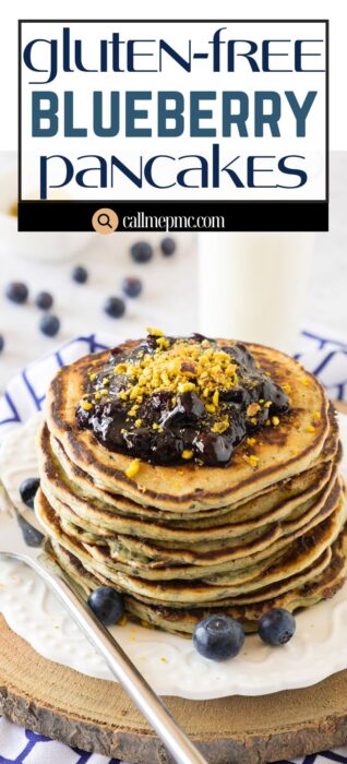 A stack of gluten-free blueberry pancakes topped with blueberry sauce and citrus zest, garnished with fresh blueberries, sits on a wooden board with a fork and a glass of milk in the background.