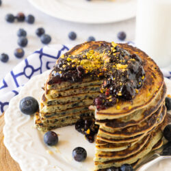 A stack of pancakes topped with dark berry sauce and crushed pistachios, served on a white plate with fresh blueberries. A fork and a glass of milk are nearby, with more pancakes in the background.