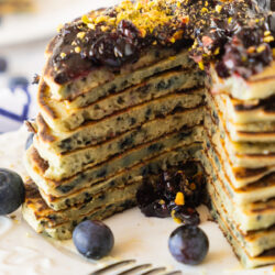 A tall stack of blueberry pancakes with a slice cut out, topped with blueberry compote and golden crumbs, served on a white plate with fresh blueberries and a fork.