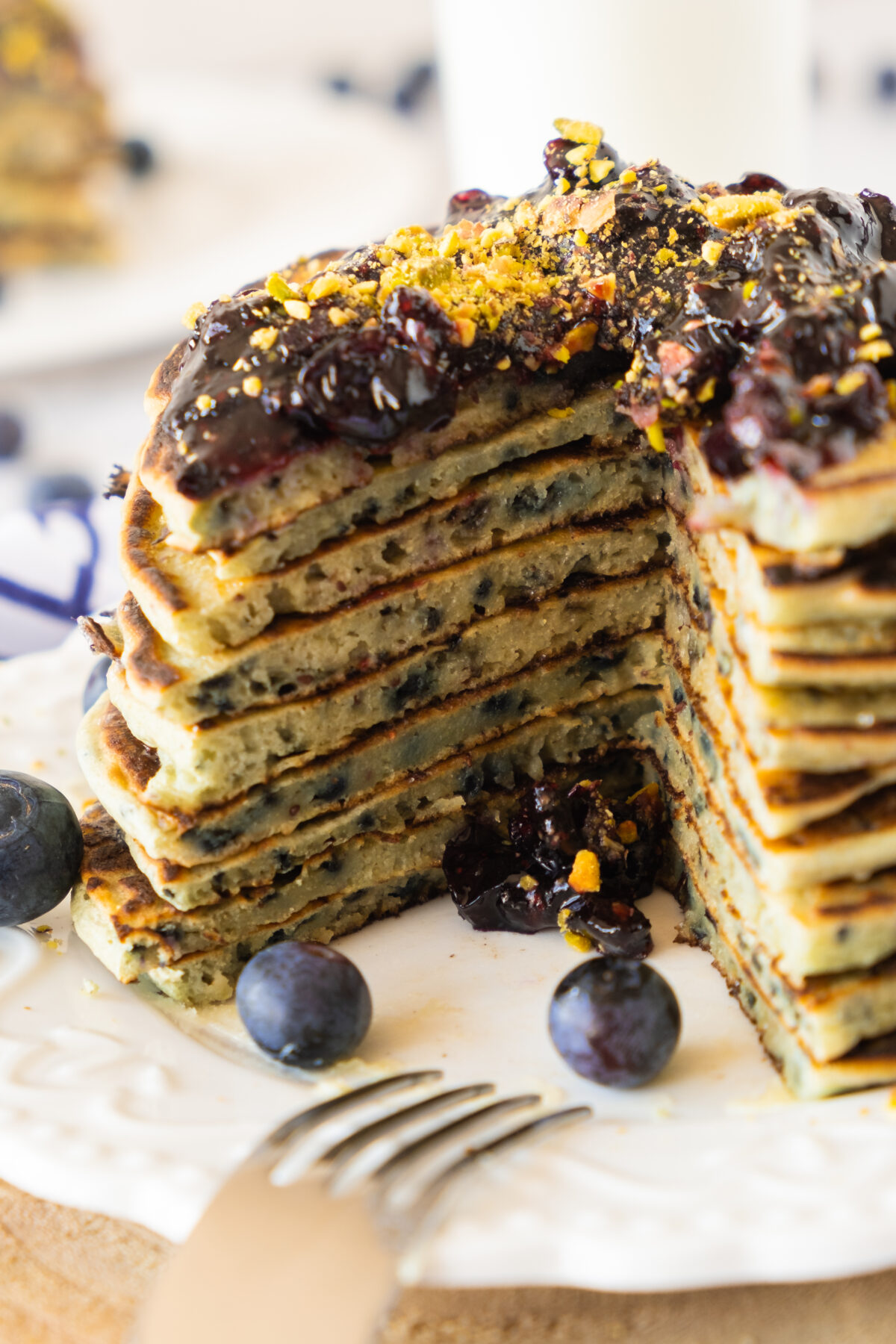 A tall stack of Blueberry pancakes with blueberry jam with a slice cut out served on a white plate with fresh blueberries and a fork.