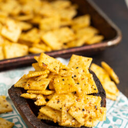 A wooden bowl filled with seasoned square crackers sits on a patterned cloth. In the background, more crackers are scattered on a baking tray and the surface below.
