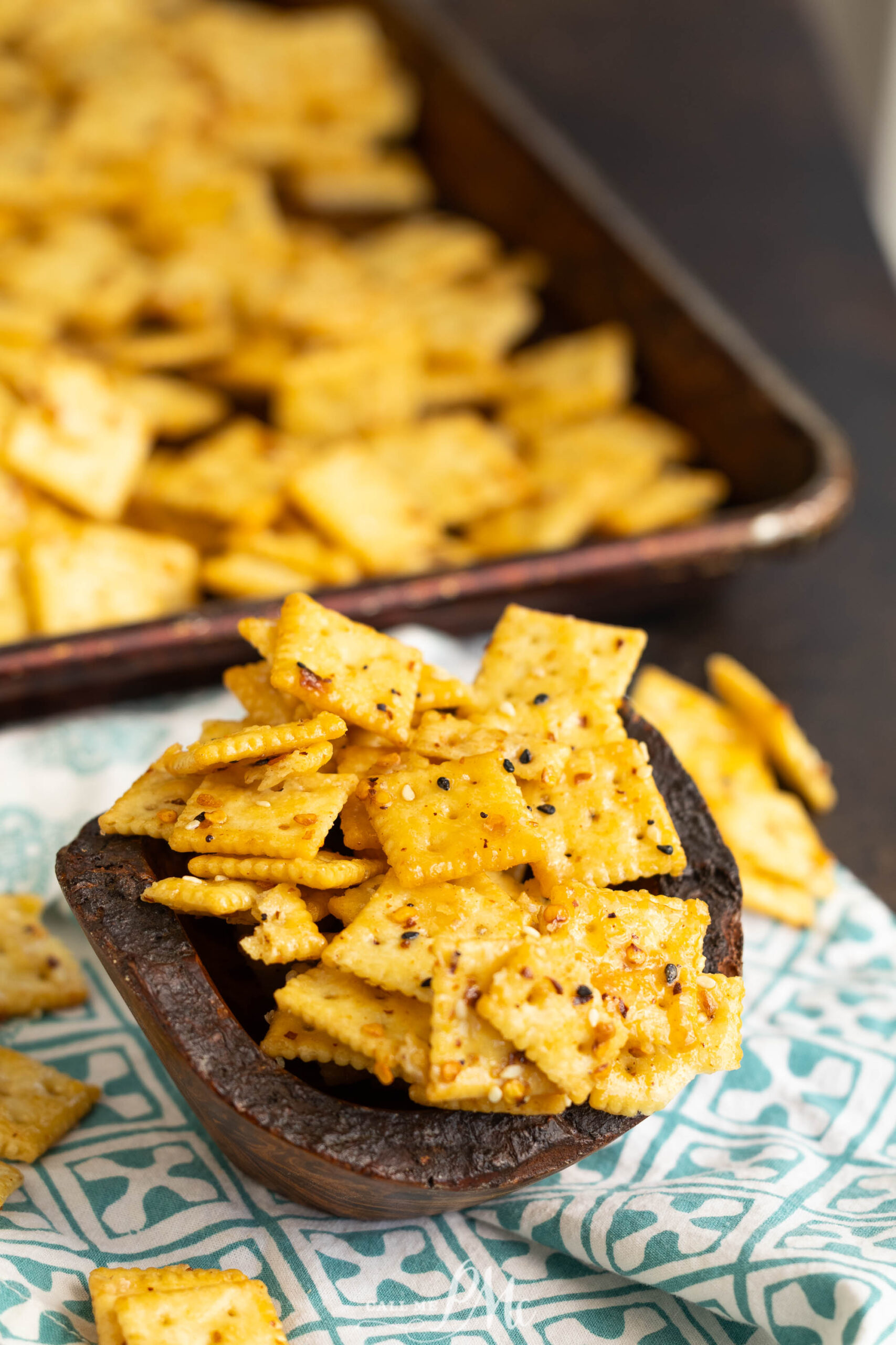 A wooden bowl filled with seasoned square crackers sits on a patterned cloth. In the background, more crackers are scattered on a baking tray and the surface below.