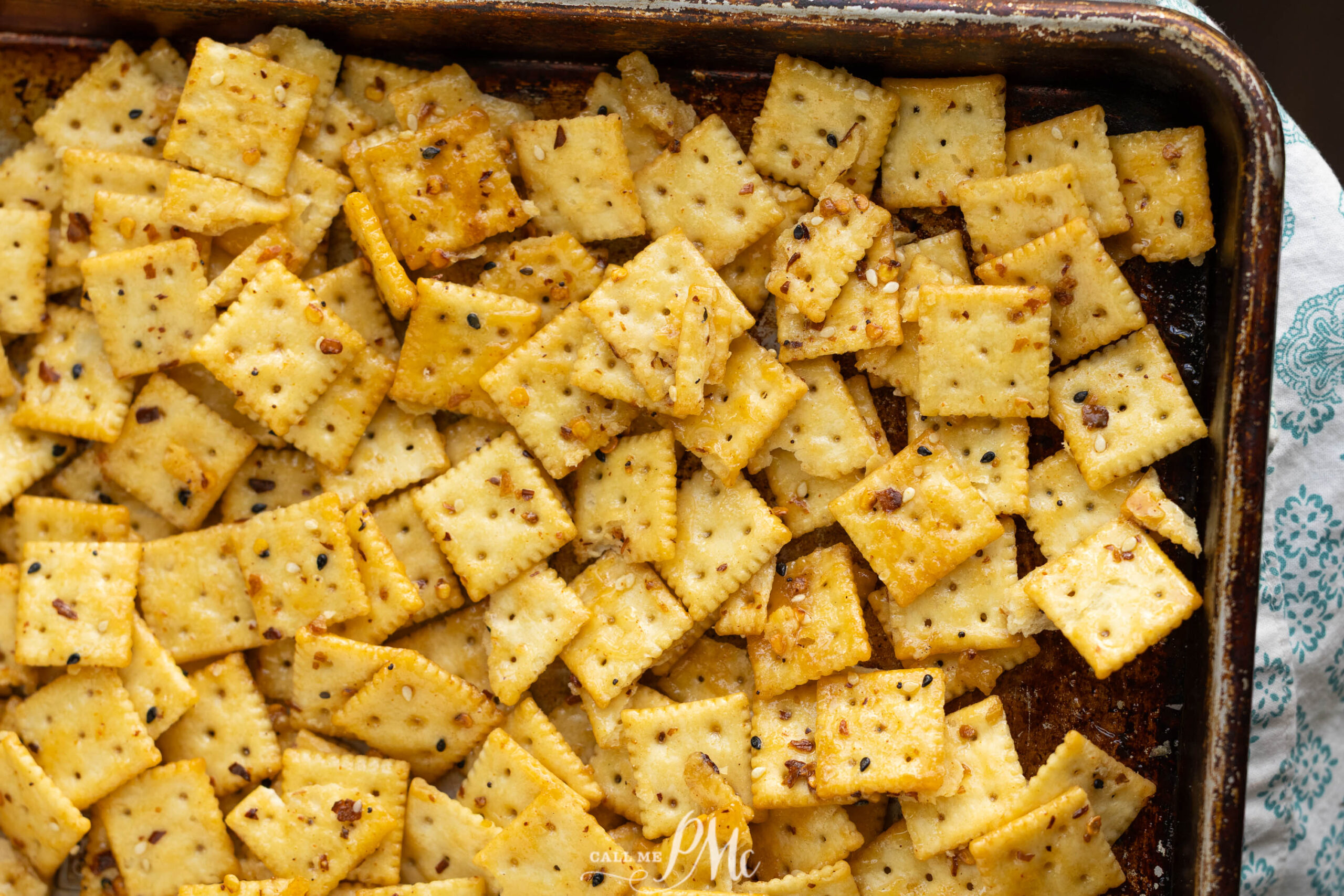 A baking sheet filled with seasoned square cheese crackers, sprinkled with spices and herbs, some slightly browned at the edges.