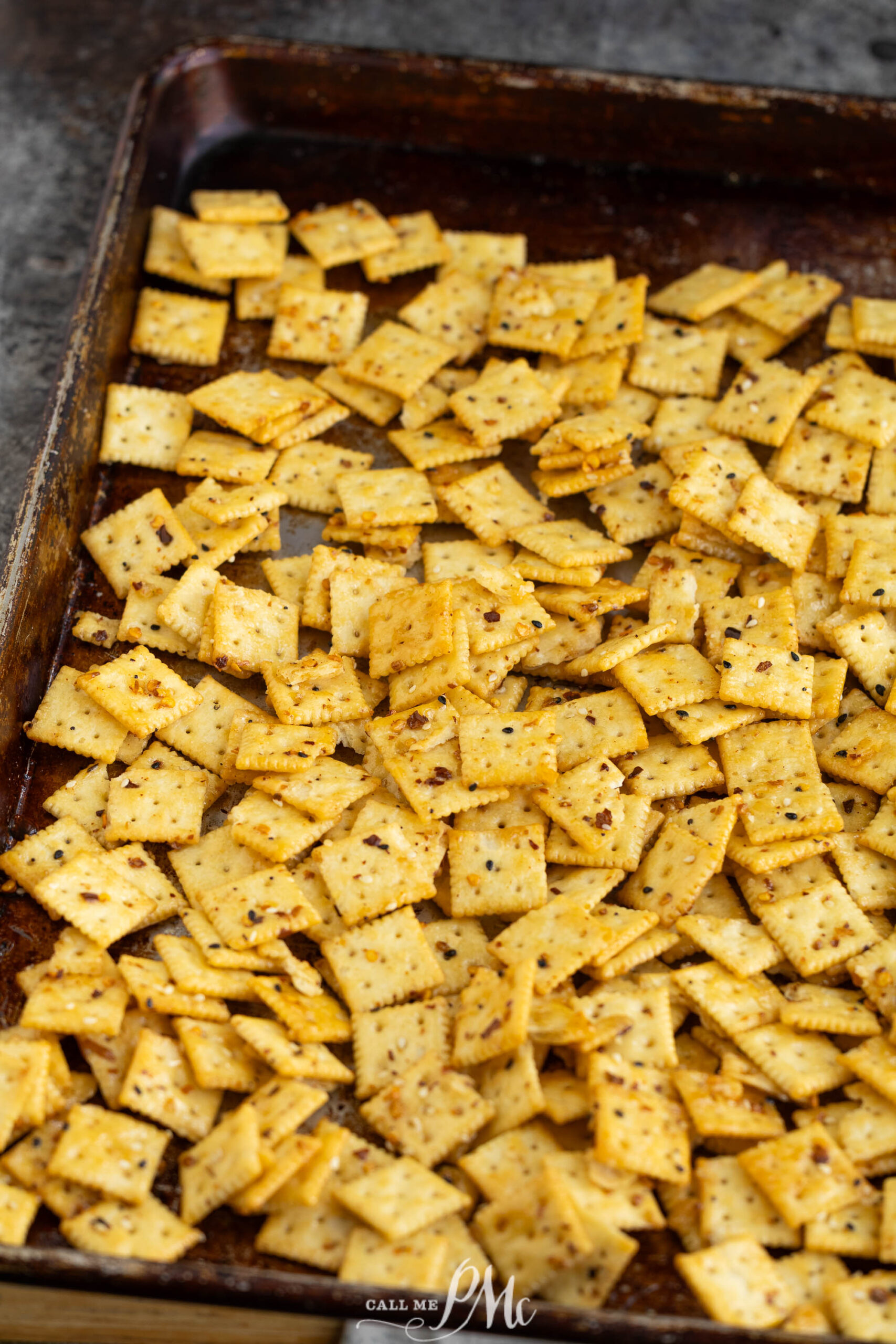 Seasoned square crackers are spread out on a dark baking sheet, ready for baking. The crackers are lightly golden and sprinkled with visible herbs and spices.