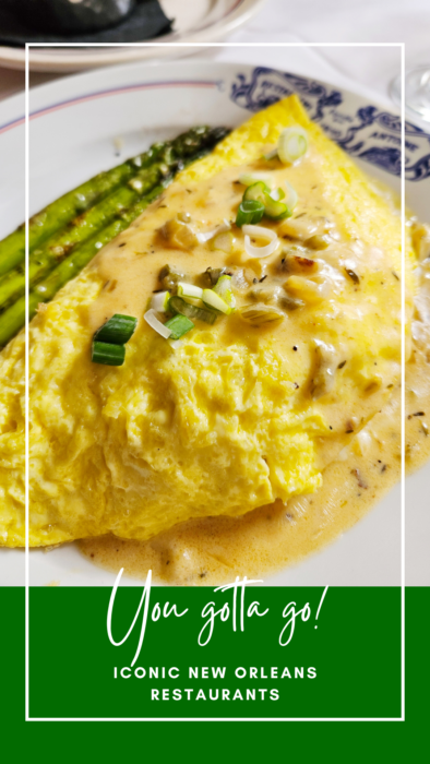 A close-up of a cheesy omelet topped with sliced green onions and creamy sauce, served with asparagus on a white plate. Text overlay reads: "You gotta go! Iconic New Orleans Restaurants.