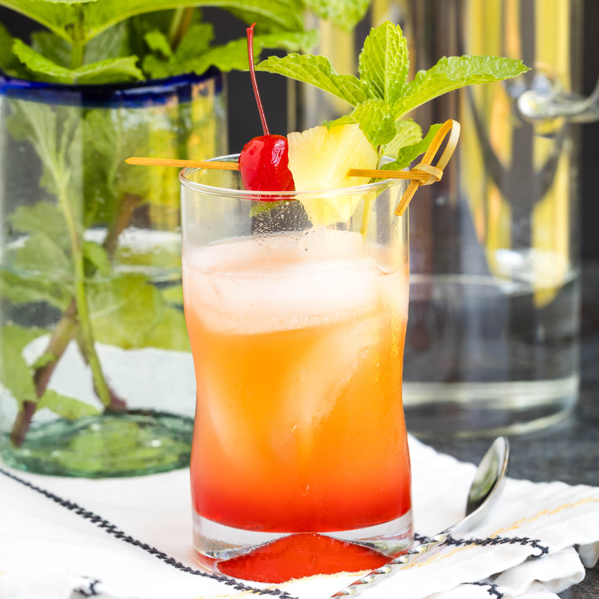 A colorful tropical cocktail with ice, garnished with a cherry, pineapple slice, and mint sprig, sits on a white napkin. Fresh mint leaves and glassware are in the background.