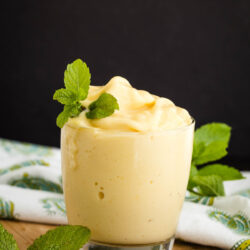 A glass filled with creamy, yellow pineapple Dole Whip topped with fresh mint leaves, placed on a wooden surface with a patterned cloth and mint sprigs in the background.