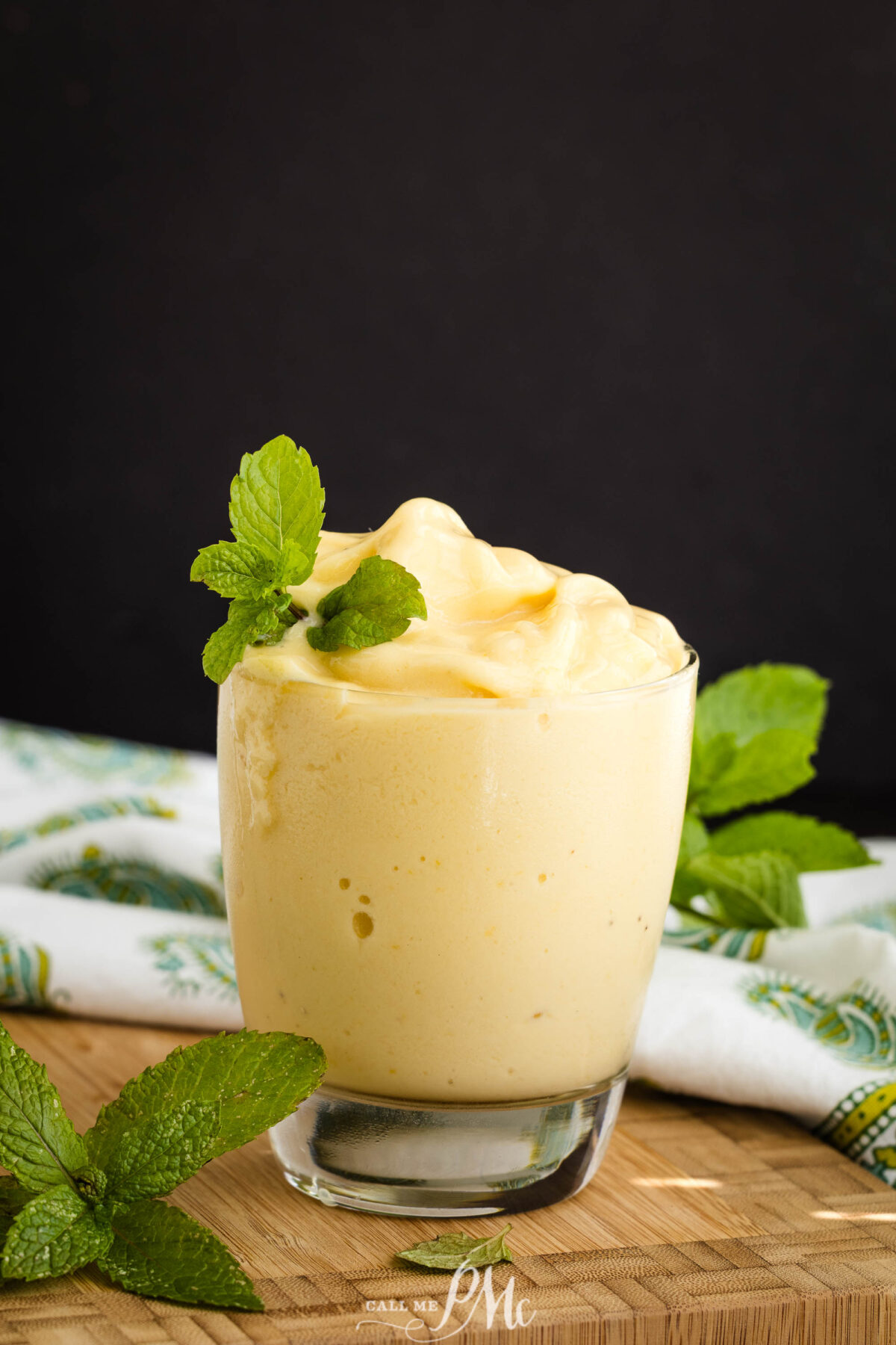 A glass filled with Dairy-free pineapple coconut smoothie for breakfast topped with fresh mint leaves, placed on a wooden surface with a patterned cloth and mint sprigs in the background.